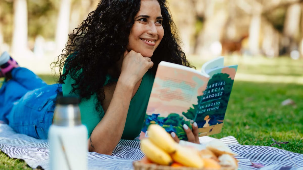Smiling woman reading a book at a picnic in a sunny park setting.